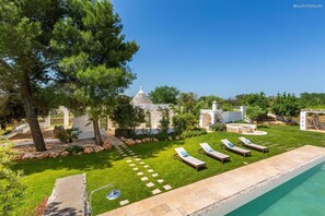 Un área junto a la piscina con sillones de descanso, un sendero de piedra y un pequeño edificio con techo cónico.