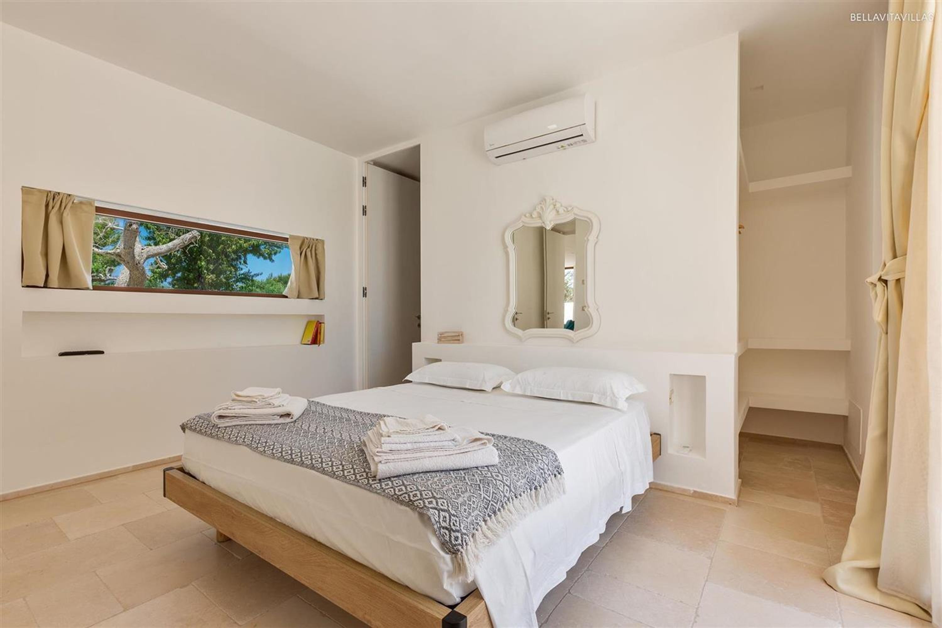 A bedroom with a bed, a window with curtains, a mirror, and an air conditioning unit.