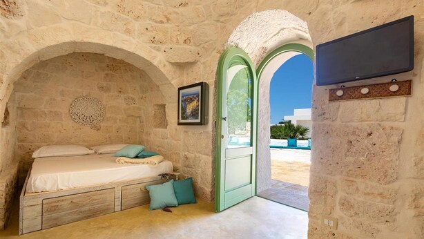 Quarto com paredes de pedra, uma cama, uma porta verde e uma televisão na parede.