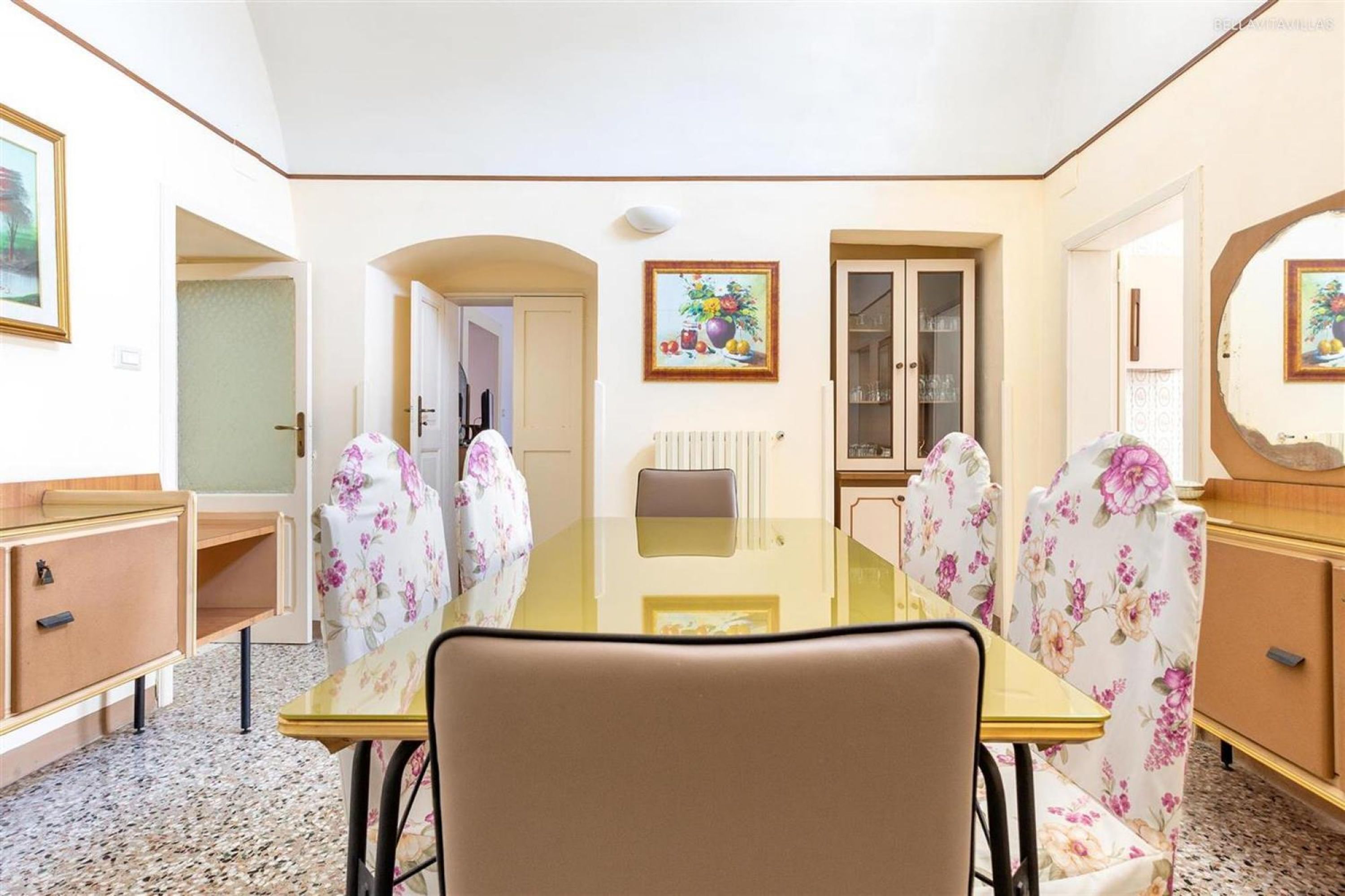 Een eetgedeelte met een glazen tafel, bloemenstoelen, een houten kast en een deur naar een andere kamer.