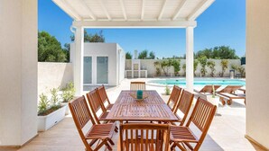 A covered outdoor area with a pool, wooden furniture, and a white building in the background. - 2055 Villa San Teodoro (Avetrana)