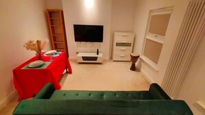 Ein gemütlicher Wohnbereich mit einem grünen Sofa, einem Esstisch mit roter Tischdecke, einem Flachbildfernseher, einer kleinen Kommode und einem Fenster mit Vorhängen.