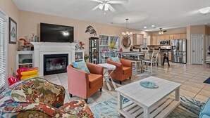 A spacious living room with a fireplace, orange armchairs, a white coffee table, and a TV. The kitchen is visible in the background with modern appliances. - Surfside Beach Family Retreat w/ Private Pool & 5BRs (Surfside Beach)