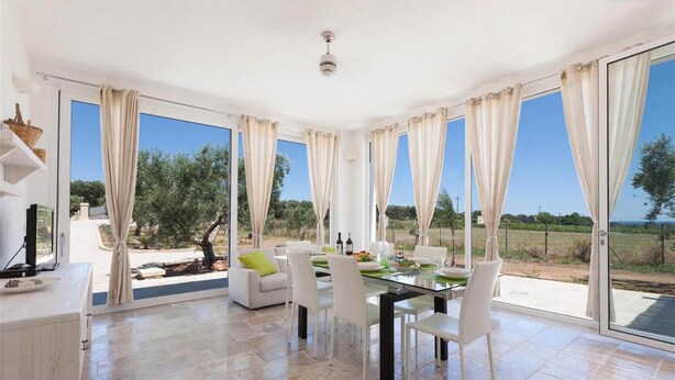 A spacious dining area with a glass table and chairs, large windows with curtains, and a view of the outdoors. - 496 La Serrazza in Pescoluse Posto Vecchio (Salve)