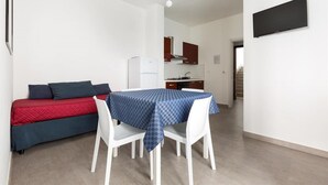 A compact living space with a dining table, white chairs, a sofa with red and blue bedding, and a kitchen area in the background. - 1298 Versomare - Monolocale Lu Ientu (Porto Cesareo)