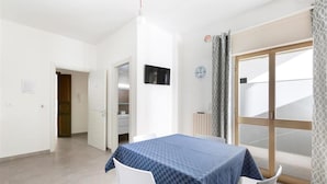 A room with a blue checkered table, white chairs, a TV on the wall, and a sliding glass door with patterned curtains. - 1298 Versomare - Monolocale Lu Ientu (Porto Cesareo)