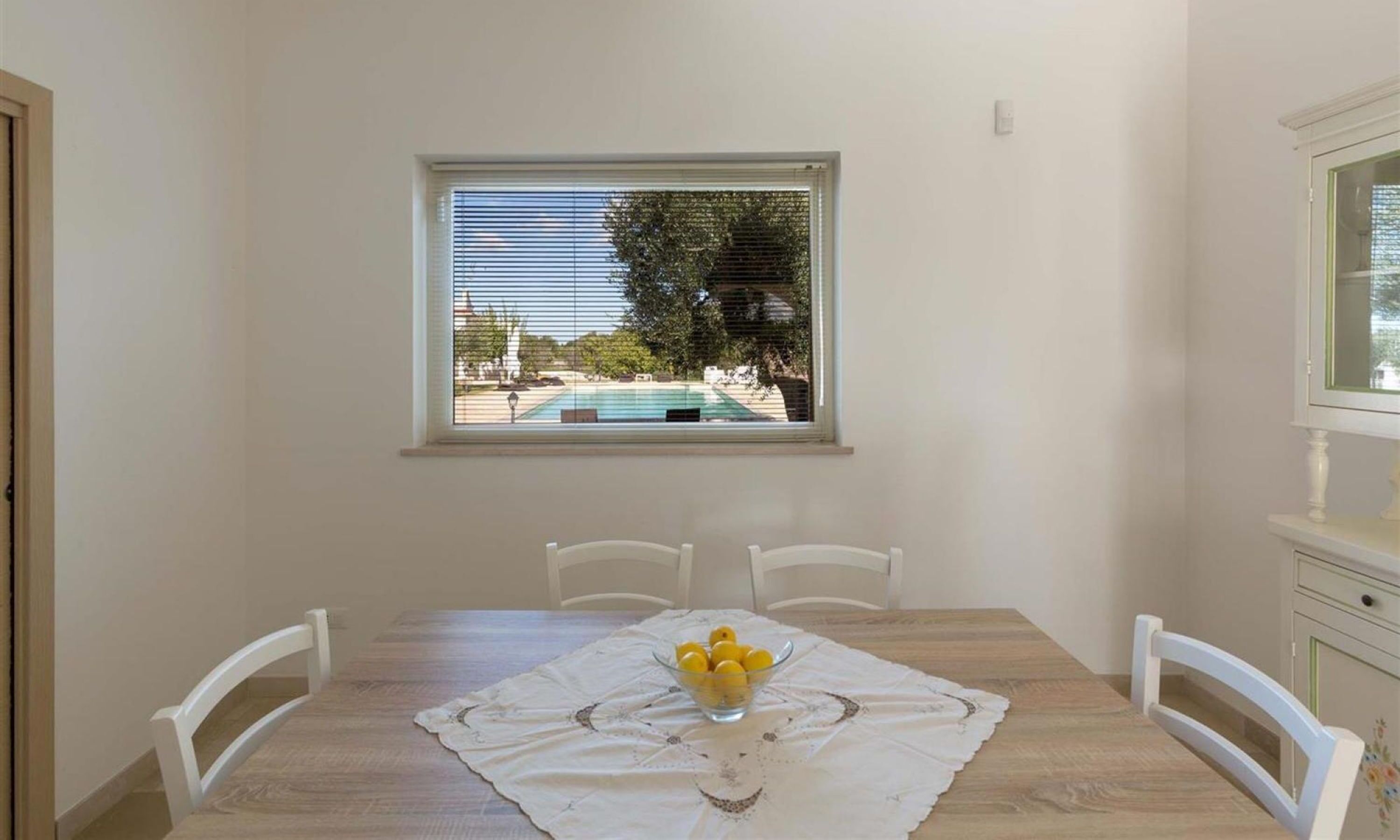 A dining area with a wooden table, white chairs, and a window with a view of a swimming pool.