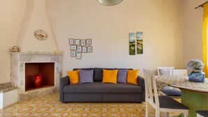 Ein Wohnzimmer mit grauem Sofa, gelben und violetten Kissen, einem Steinkamin, einem gemusterten Boden und einem Essbereich mit Tisch und Stühlen.