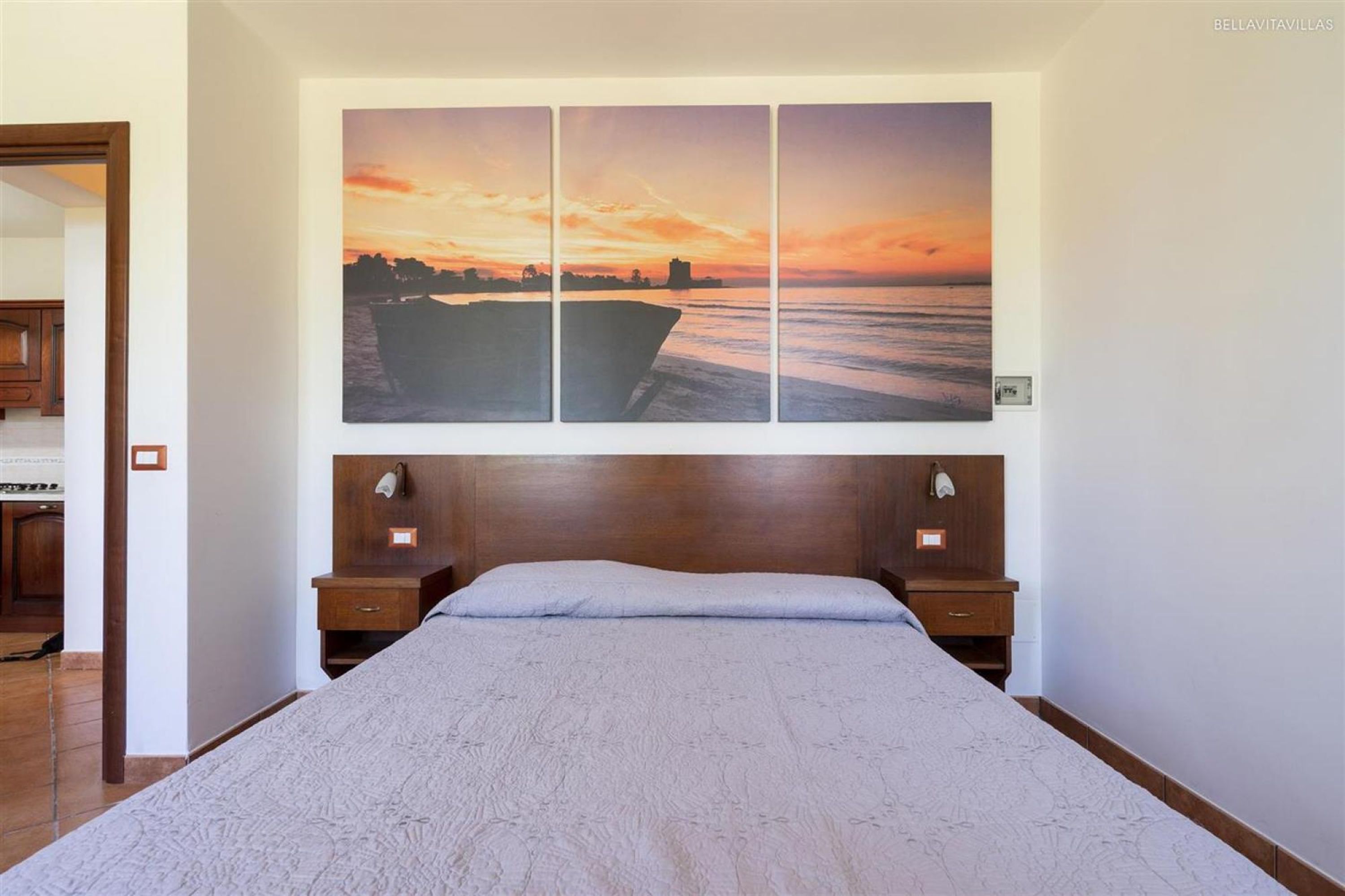 Une chambre à coucher avec un grand lit, une tête de lit en bois, deux tables de chevet et un tableau accroché au mur représentant un coucher de soleil sur la mer.