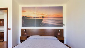 Un dormitorio con una cama grande, cabecera de madera, dos mesitas de noche y un cuadro colgado en la pared que muestra un atardecer sobre el mar.
