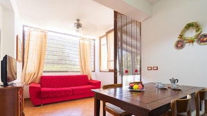 Una sala de estar con un sofá rojo, una mesa de comedor de madera y sillas. Hay un televisor sobre un mueble de madera, y una ventana con cortinas.