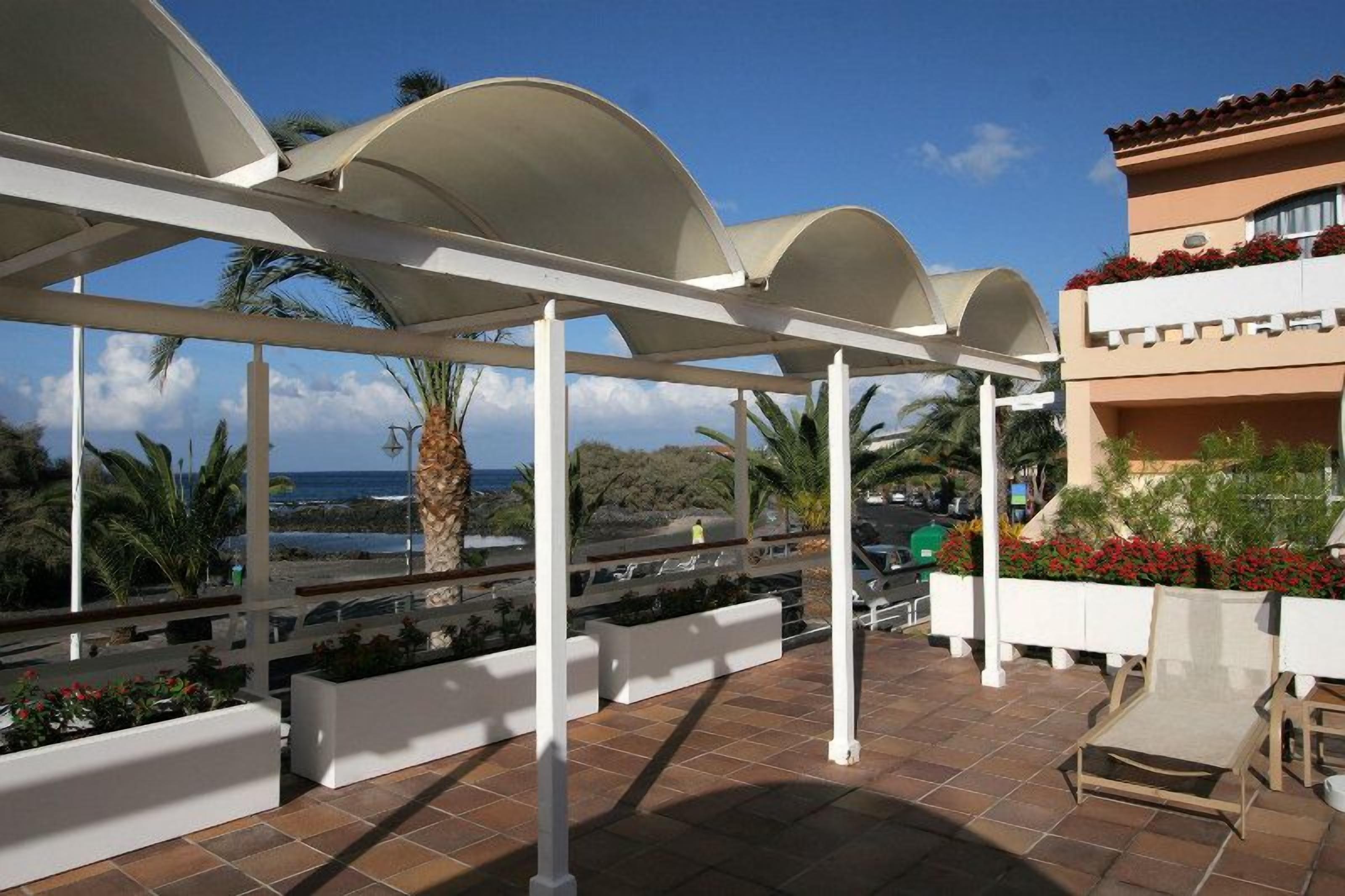 A covered outdoor area with a view of the sea, white columns, and a building with red flowers.