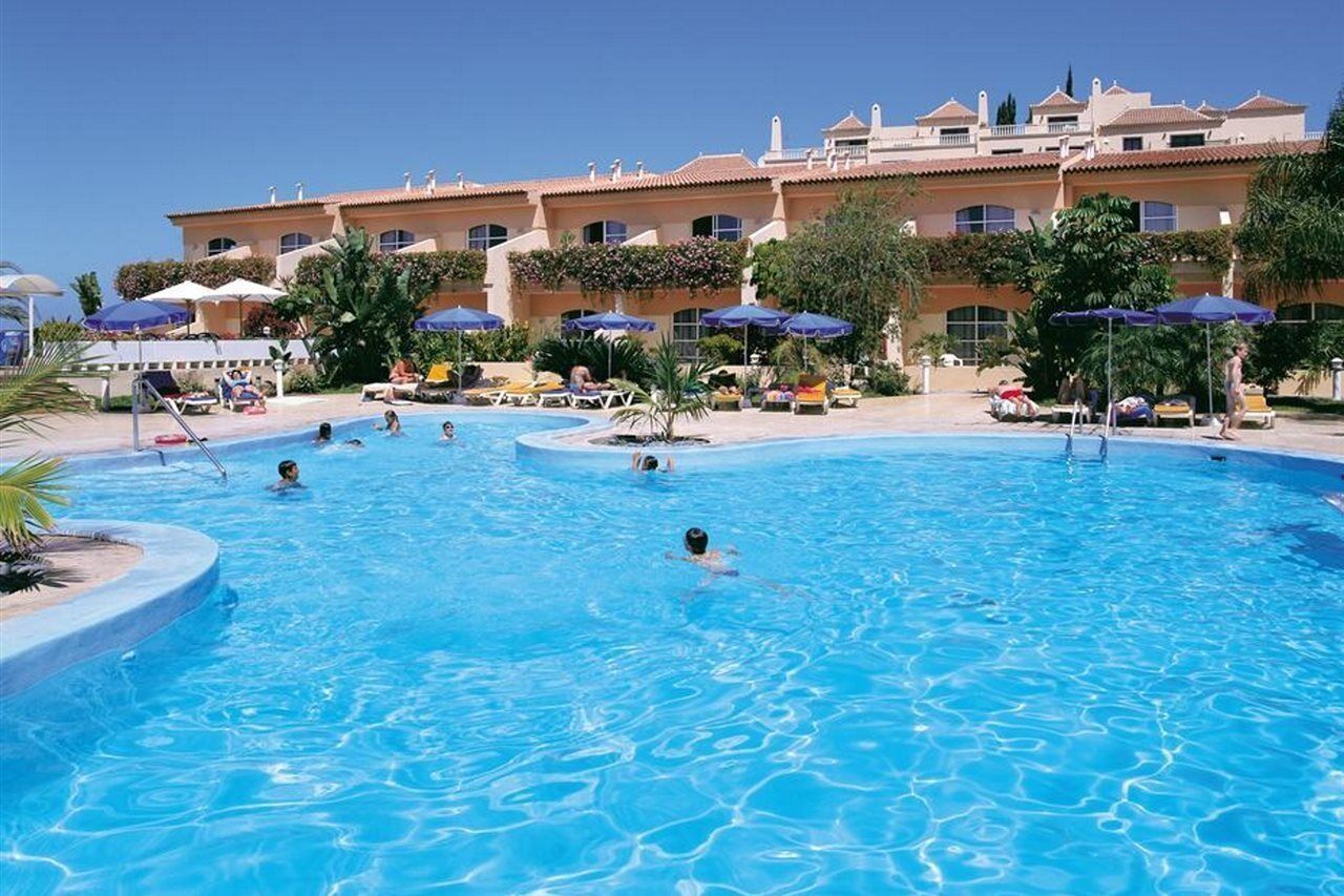 A swimming pool with multiple lanes, surrounded by lounge chairs and umbrellas, and a building with a tiled roof in the background.