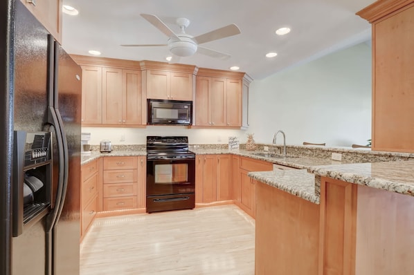 A kitchen with wooden cabinets, a microwave, a stove, a ceiling fan, and a granite countertop.