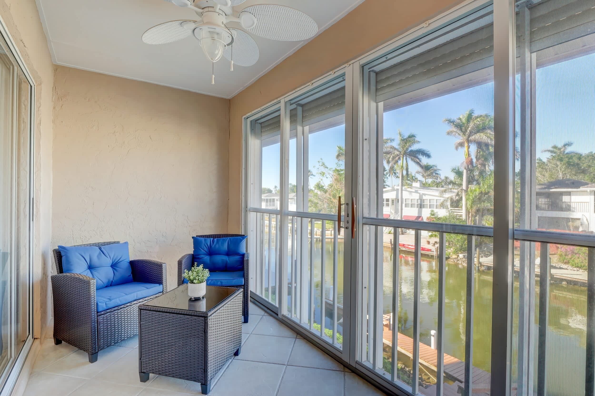 A balcony with wicker furniture, a ceiling fan, and a view of a body of water and palm trees.