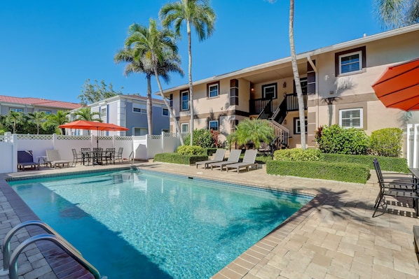 A swimming pool with lounge chairs and umbrellas, surrounded by palm trees and a building with a staircase.
