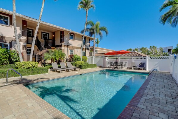 A swimming pool with a red umbrella, lounge chairs, and palm trees.
