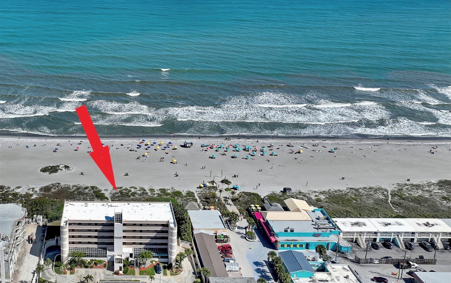 An aerial view of a coastal area with a beach, a parking lot, and a building with multiple floors and a flat roof.