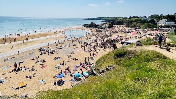 Una playa concurrida con gente y tiendas de campaña, cielo despejado y vista al mar.