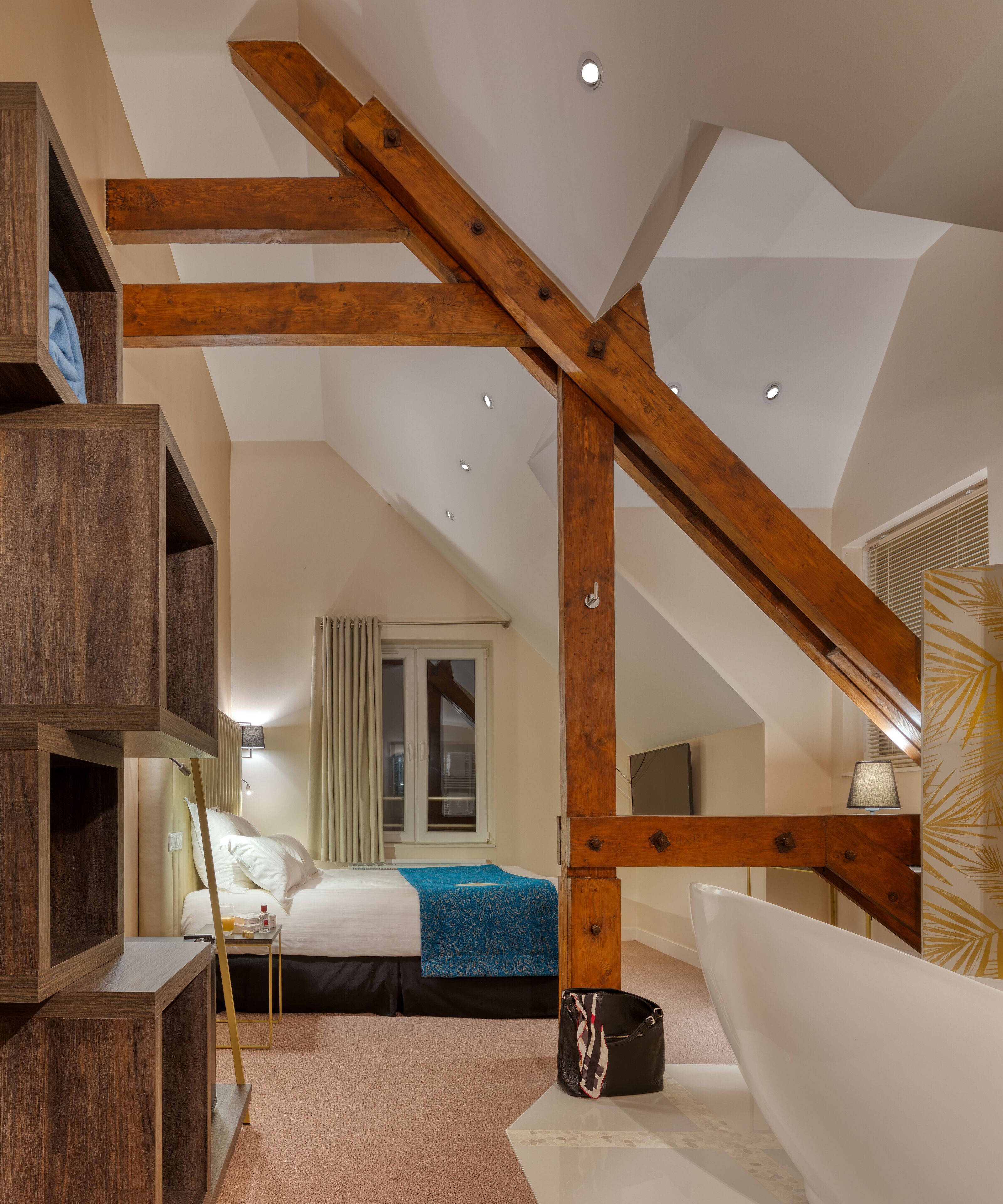 A bedroom with a sloped ceiling, wooden beams, a bed with a blue bedspread, a bedside table, a lamp, and a large window with curtains.