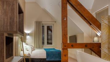 A bedroom with a sloped ceiling, wooden beams, a bed with a blue bedspread, a bedside table, a lamp, and a large window with curtains.