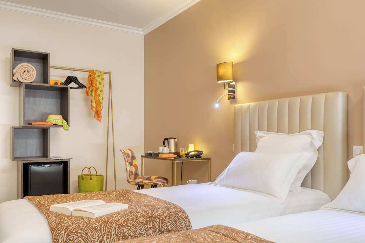 A hotel room with two beds, a bedside table with a lamp, a chair, and a shelf with towels and a hat.