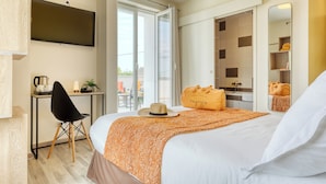 Habitación de hotel con cama, escritorio con silla, televisor, baño con ducha y balcón con mobiliario exterior.