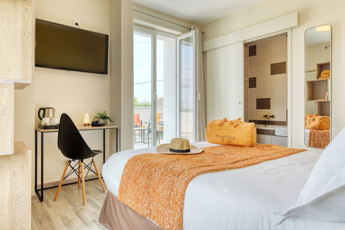 A hotel room with a bed, a desk with a chair, a television, a bathroom with a shower, and a balcony with outdoor seating.