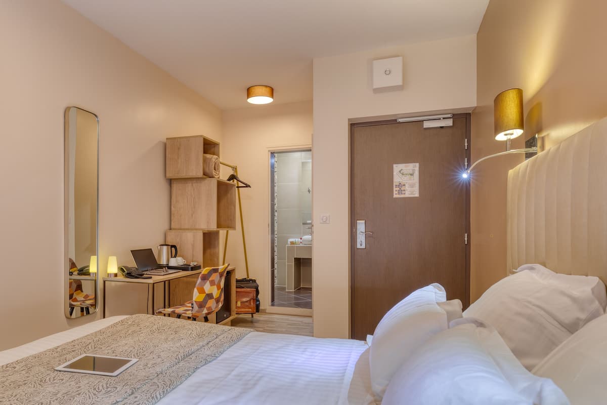 A hotel room with a bed, bedside table, lamp, desk with a chair, and a mirror.