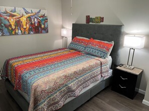 A bedroom with a patterned bedspread, a grey headboard, two bedside lamps, a small bedside table, and a wall-mounted artwork.