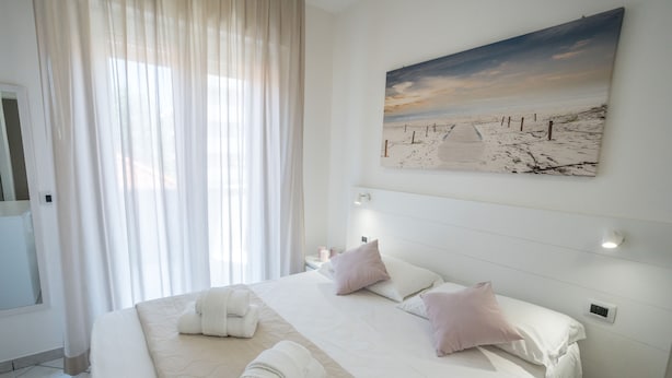 Una habitación de hotel con una cama grande, un cuadro de una playa y una ventana con cortinas translúcidas.