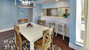 A dining area with a white table, wooden chairs, and a chandelier. There is a kitchen area with a counter and stools.
