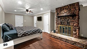 Ein Schlafzimmer mit einem Bett, einem Deckenventilator, einem Kamin aus Stein und einem an der Wand befestigten Fernseher.
