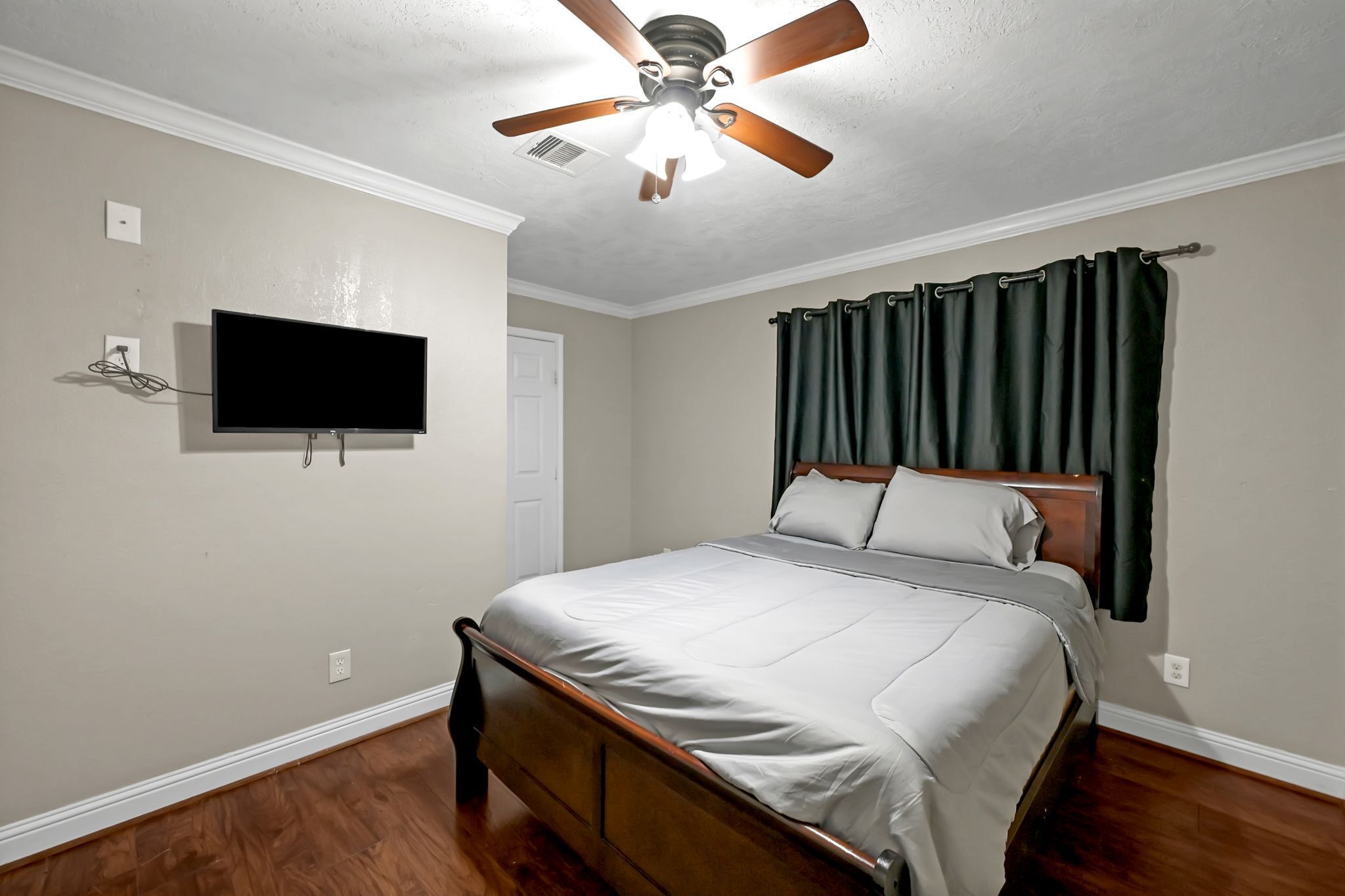 A bedroom with a bed, a ceiling fan, a television mounted on the wall, and a door.
