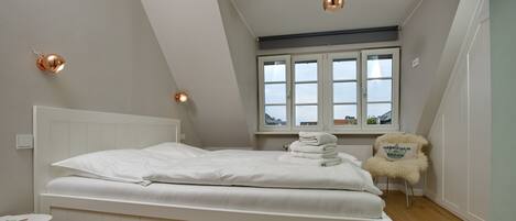 A bedroom with a white bed, a small round table, a chair with a cushion, and a window with a view of the outside.