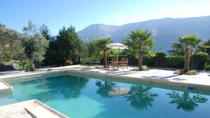 A clear swimming pool with a view of mountains in the background, surrounded by palm trees and a patio area with chairs and an umbrella. - Cortijo for 6 with Large Swimming Pool in 36 Acres of Land in Sierra Nevada Park (Granada)