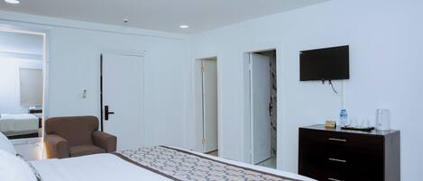 Habitación de hotel con cama, una silla, cómoda, televisor y puerta que da a otra habitación.
