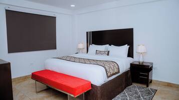 A hotel room with a bed, bedside tables, a red bench, a patterned rug, and a large window.