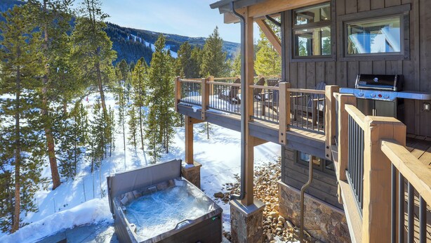 A wooden deck with a hot tub, outdoor seating, and a grill area overlooking a snowy landscape with trees and mountains. - Custom Mountain Retreat – Hot Tub & Summer Views (Keystone)