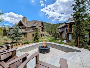 Una cabaña con chimenea de piedra, mobiliario exterior y montañas al fondo.