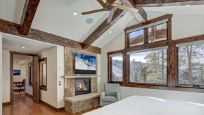 A bedroom with a stone fireplace, a large bed, a television, and a window with a view of trees. - Custom Mountain Retreat – Hot Tub & Summer Views (Keystone)