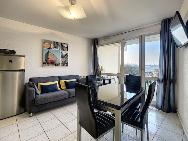 A modern living room with a sofa, dining table, chairs, a refrigerator, and a large window offering a city view.