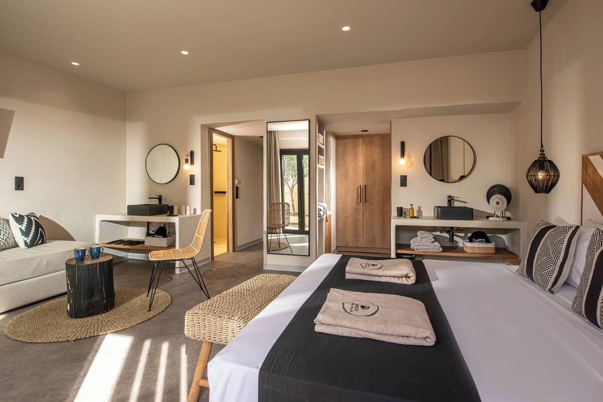 A modern hotel room with a large bed, a sitting area with a sofa and chair, a small desk, and a wardrobe.