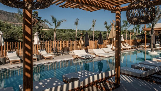 A pool area with lounge chairs and umbrellas, surrounded by wooden fences and palm trees. - Indigo Inn Casa - Adults Only (Hersonissos)
