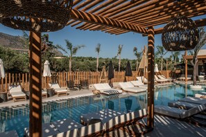 Zona de piscina con sillas de descanso y sombrillas, rodeada de cercas de madera y palmeras.