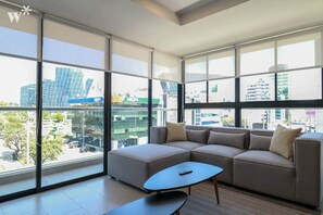 A modern living room with a sectional sofa, a coffee table, and large windows offering a city view. - w* | Sleek 1BR in San Isidro (San Isidro)