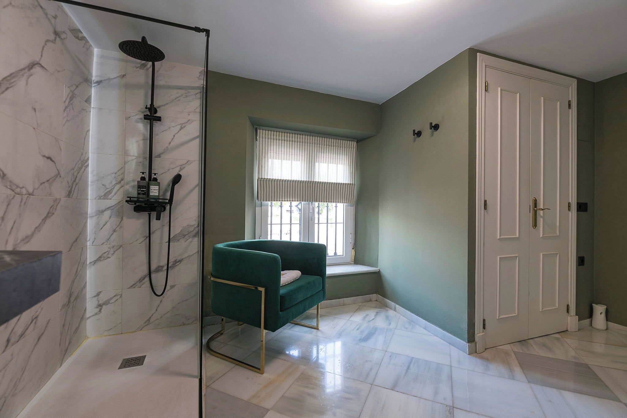 A bathroom with a shower, a bench, a window with curtains, and a door.
