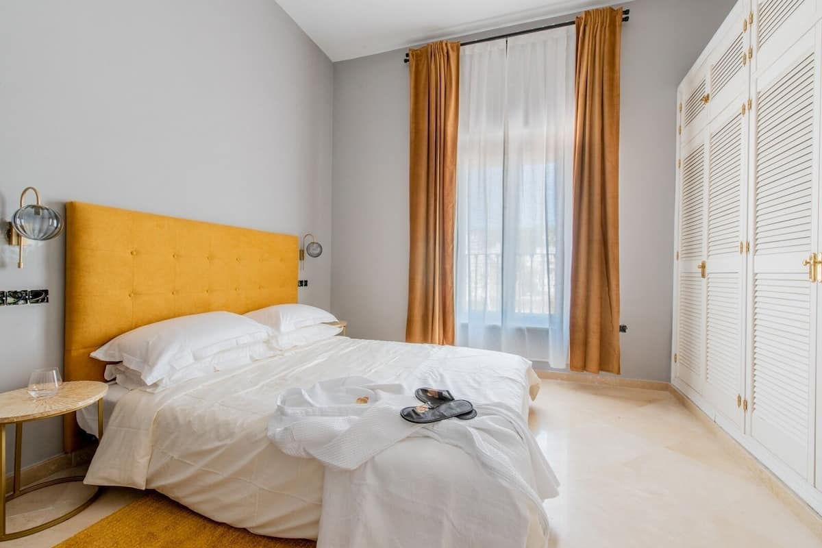 Quarto com cama, mesa de cabeceira, guarda-roupa e janela com cortinas.