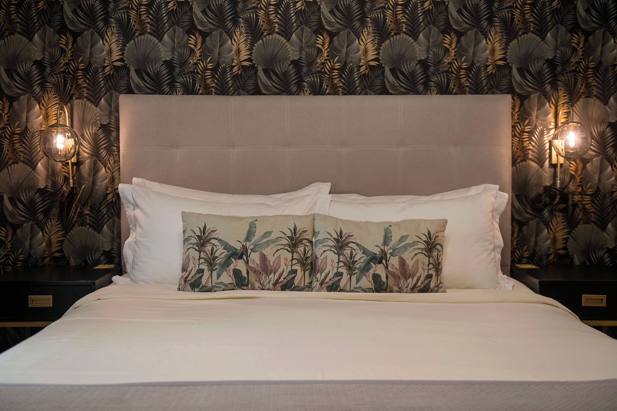 A neatly made bed with white linens and a single decorative pillow, set against a wall with a leaf-patterned wallpaper and two wall-mounted lamps.