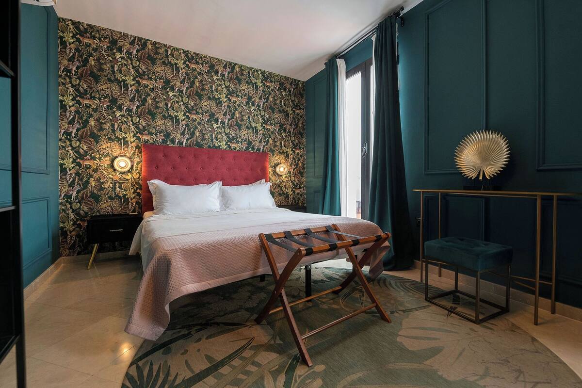 Quarto de hotel com uma cama grande, uma mesa de cabeceira, uma pequena mesa com abajur e um objeto decorativo na parede.